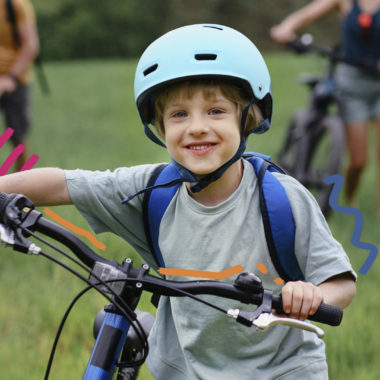 Foto de um menino de pele clara vestindo camiseta cinza e mochila de alças azuis nas costas. Ele está em pé ao lado da bicicleta e usa capacete, ambos azuis.