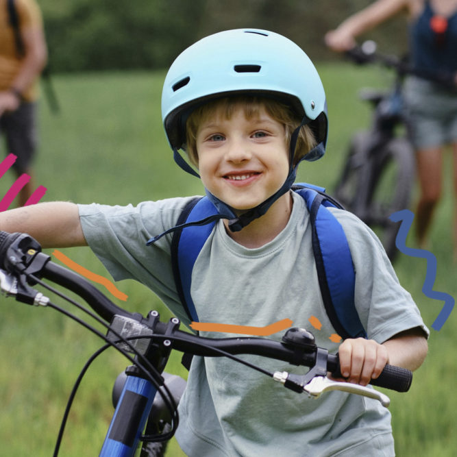 Foto de um menino de pele clara vestindo camiseta cinza e mochila de alças azuis nas costas. Ele está em pé ao lado da bicicleta e usa capacete, ambos azuis.