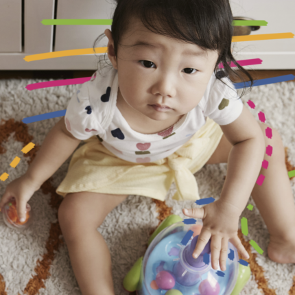 Uma menina com traços orientais está sentada em cima de um tapete e cercada de brinquedos.