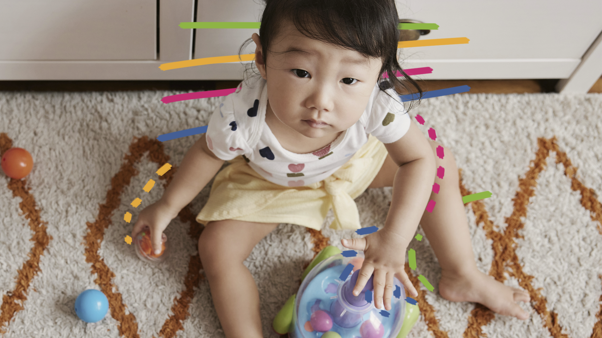 Uma menina com traços orientais está sentada em cima de um tapete e cercada de brinquedos.
