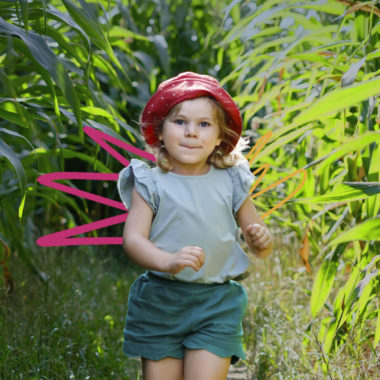 Uma menina de pele e cabelo claros está em meio a plantas, usa short, blusa e um chapéu vermelho.