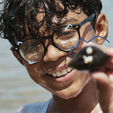 Um menino de cabelos escuros e óculos segura próximo ao rosto uma concha do mar
