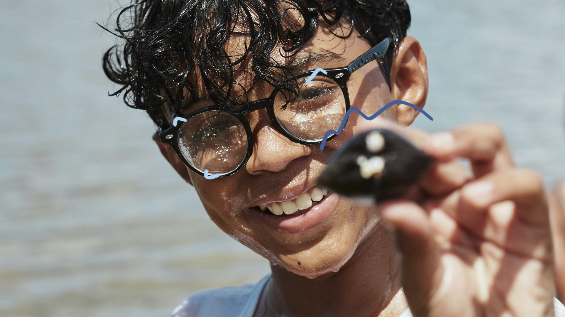 Um menino de cabelos escuros e óculos segura próximo ao rosto uma concha do mar