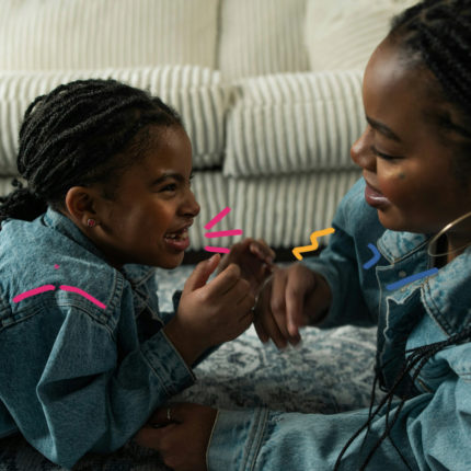 Uma mulher e uma menina, ambas negras, de tranças e usando roupas jeans, conversam no chão da sala