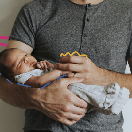 O tronco de um homem vestindo camiseta cinza escuro com botões e suas mãos que seguram um bebê recém-nascido de macacão claro.