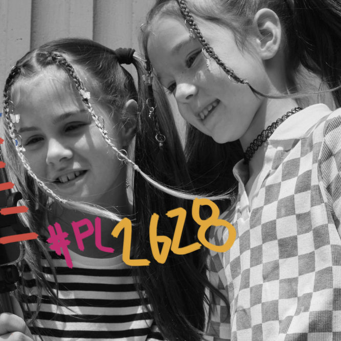 Foto em preto e branco de duas meninas olham para a tela de um celular. Uma delas está com blusa listrada e a outra quadriculada. Na imagem, há o texto: #PL2628