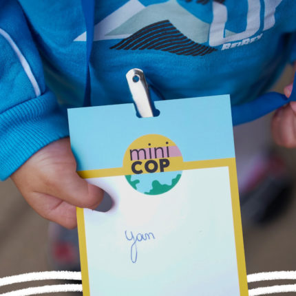 A mão de uma criança vestindo moletom azul segura um crachá com o seu nome, Yan, e um símbolo com parte do planeta terra em azul e verde escrito miniCOP