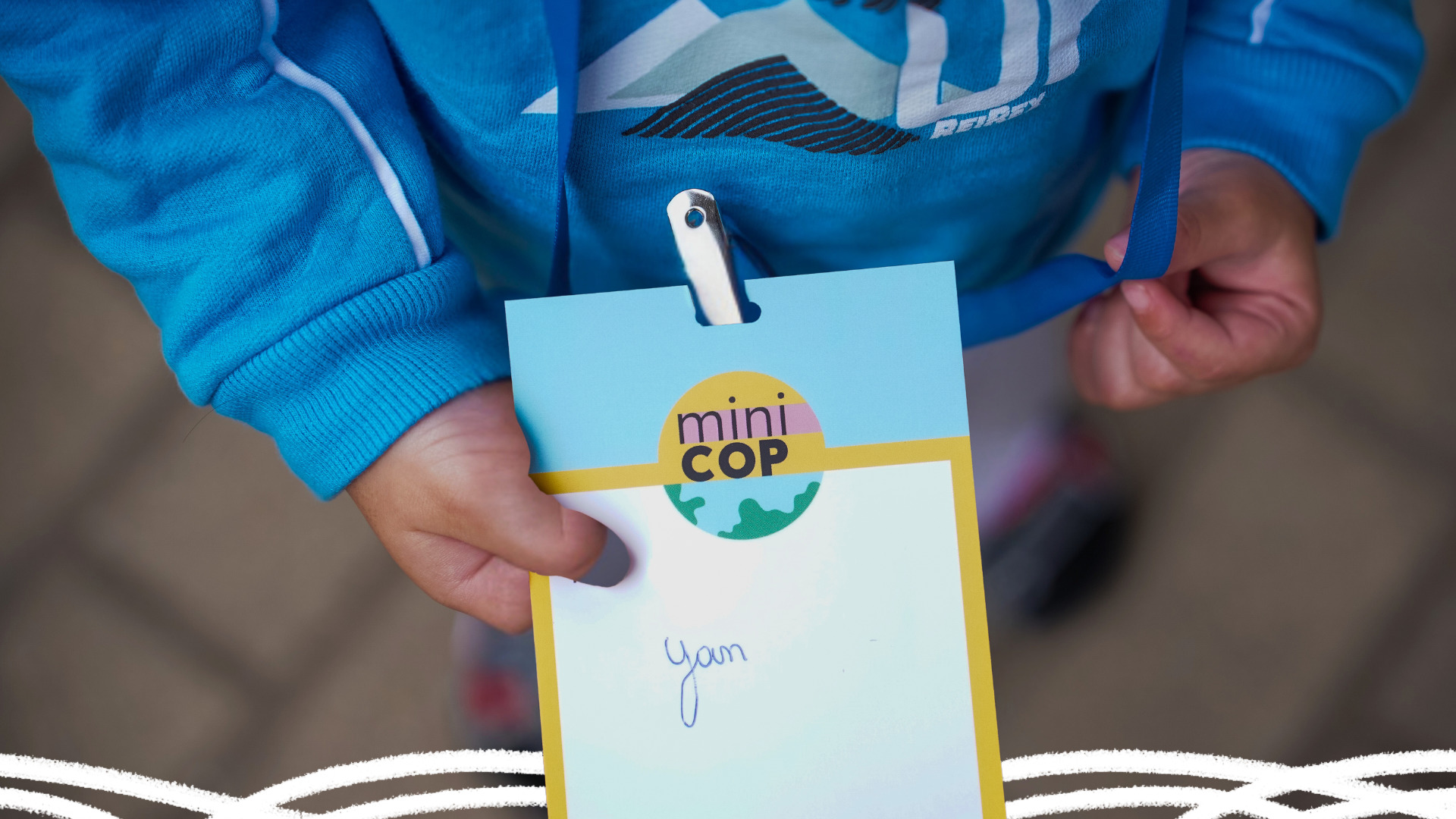 A mão de uma criança vestindo moletom azul segura um crachá com o seu nome, Yan, e um símbolo com parte do planeta terra em azul e verde escrito miniCOP