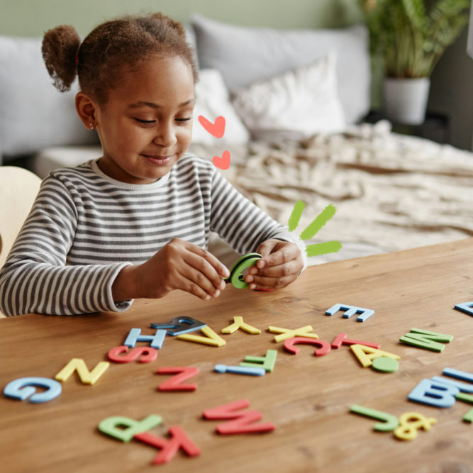 Uma menina negra de coque e blusa listrada está brincando com letras coloridas dispostas em uma mesa de madeira na sala de casa.