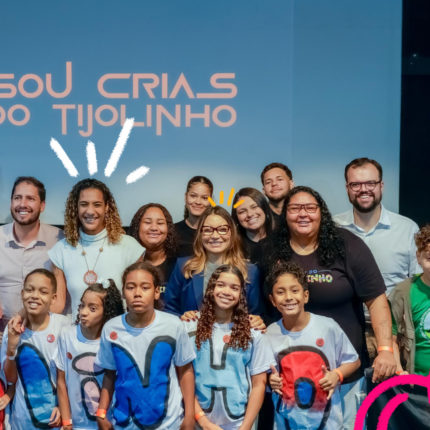 Um grupo de crianças e adultos posam para a foto em frente a uma tela azul onde está escrito Sou Crias do Tijolinho