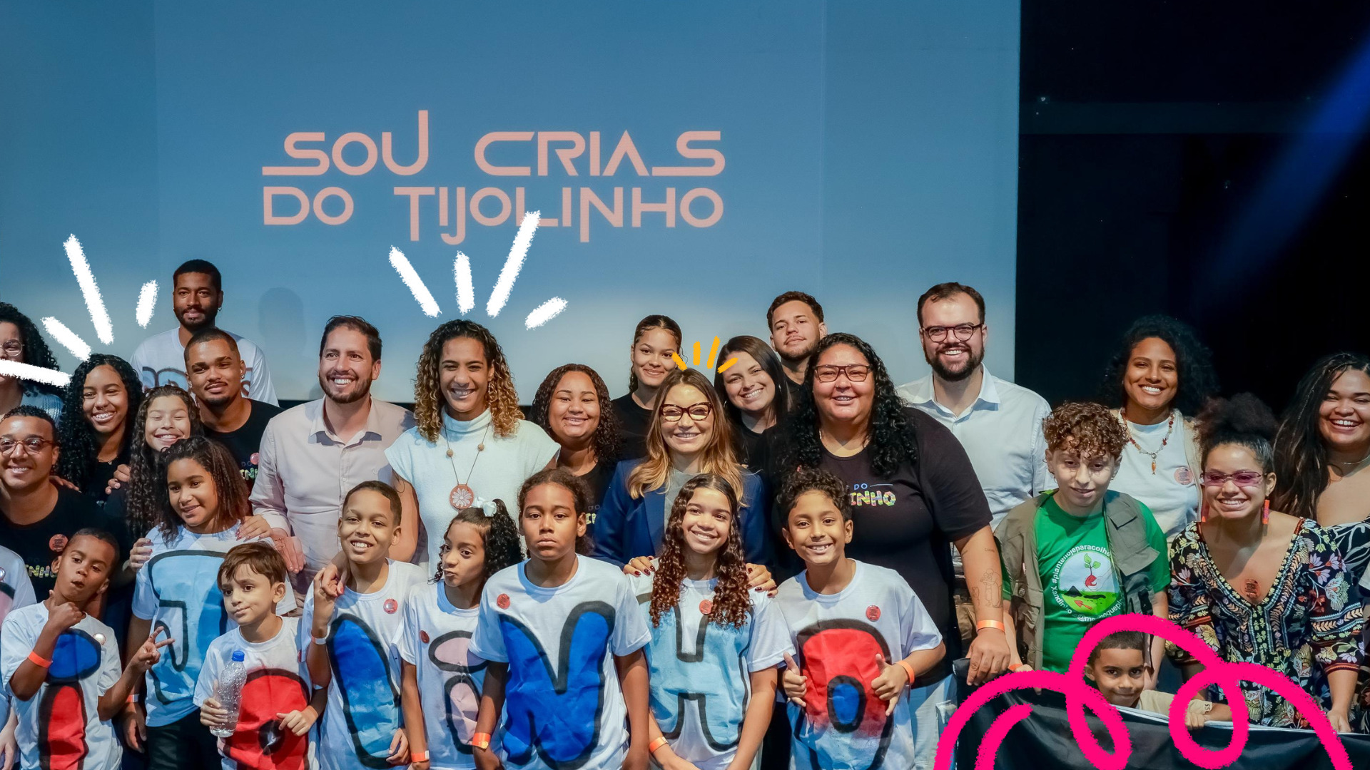 Um grupo de crianças e adultos posam para a foto em frente a uma tela azul onde está escrito Sou Crias do Tijolinho