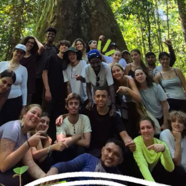 Imagem mostra projeto sobre COP30 nas escolas. Um grupo de adolescentes estão no meio de uma floresta ao redor de uma árvore.