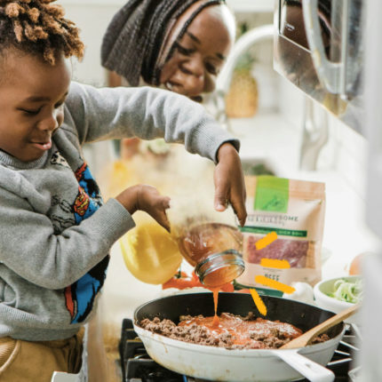 Imagem mostra uma mãe e uma criança na cozinha. Um menino negro e uma mulher negra estão preparando uma comida com panelas e talheres.