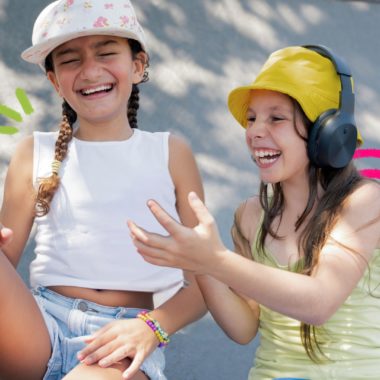 Imagem mostra duas crianças rindo. Uma veste camiseta e boné brancos, shorts jeans e meia rosa. A outra veste camiseta e boné amarelos e usa um fone de ouvido preto. Elas parecem brincar fora das telas e se divertir com isso.