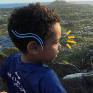 Um menino negro de perfil usa camiseta azul e está numa área em meio à natureza