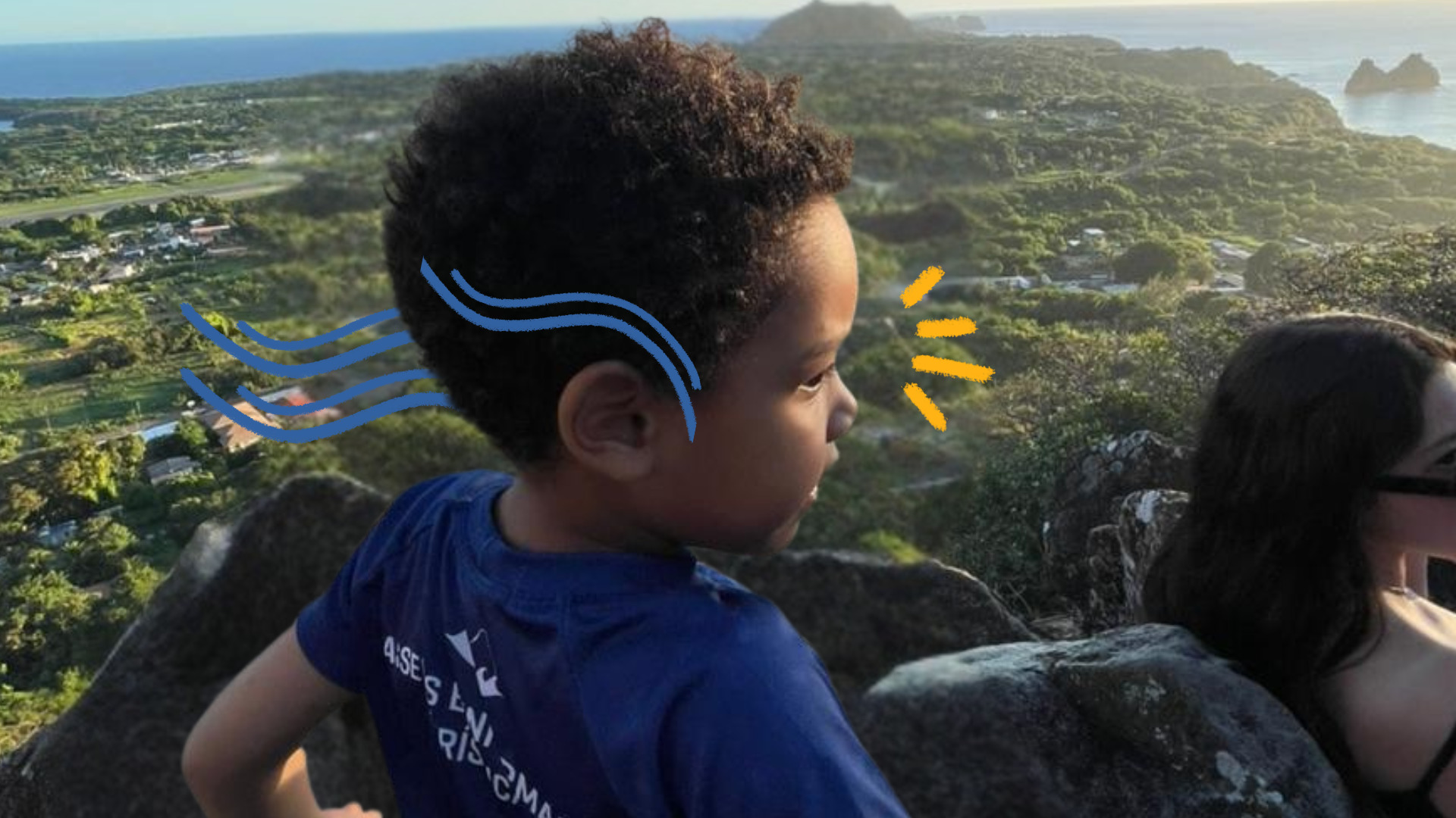 Um menino negro de perfil usa camiseta azul e está numa área em meio à natureza