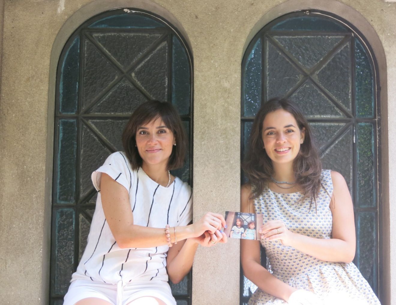 Imagem mostra as duas amigas Elisa e Talita sentadas em uma janela segurando uma foto impressa. 