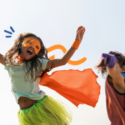 Duas crianças brincam de super-heróis. À esquerda, uma menina veste capa e máscara laranja, camiseta branca e uma saia de tule verde. Ela está levantando o braço em pose de voo. À direita, um menino usa capa azul e camiseta vermelha. Ele ergue o braço, tampando seu rosto, como se estivesse lutando.