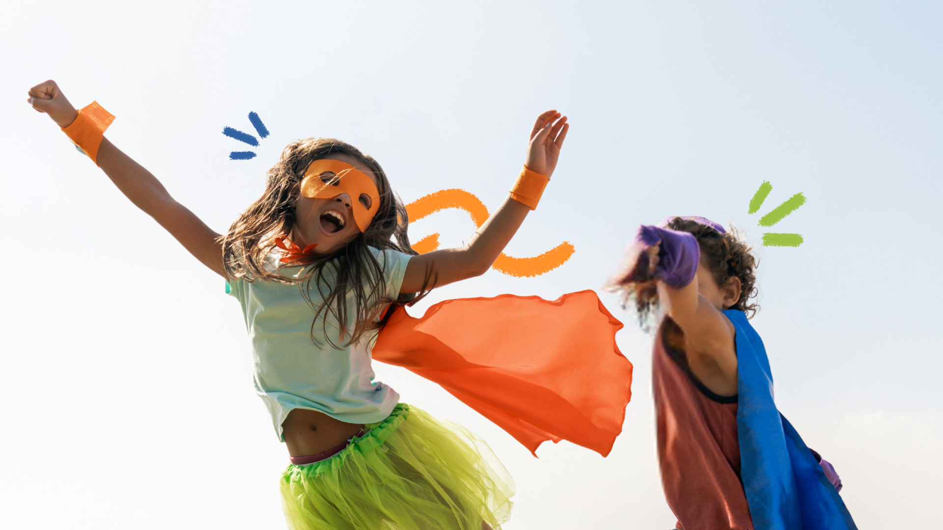 Duas crianças brincam de super-heróis. À esquerda, uma menina veste capa e máscara laranja, camiseta branca e uma saia de tule verde. Ela está levantando o braço em pose de voo. À direita, um menino usa capa azul e camiseta vermelha. Ele ergue o braço, tampando seu rosto, como se estivesse lutando.