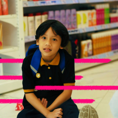 Imagem mostra menino de pele morena e cabelos pretos sentado no chão de um supermercado. Ao fundo, uma estante com produtos de higiene pessoal, como shampoo e condicionador. Ele veste uma camiseta azul marinho com gola amarela. Imagem representa como falar sobre dinheiro com as crianças.