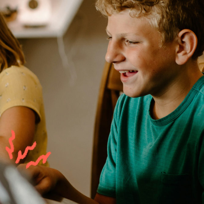 Imagem mostra um menino branco, de cabelo loiro curto e camisa verde sentando ao lado de uma menina branca de cabelos lisos e óculos. Eles estão usando computadores.