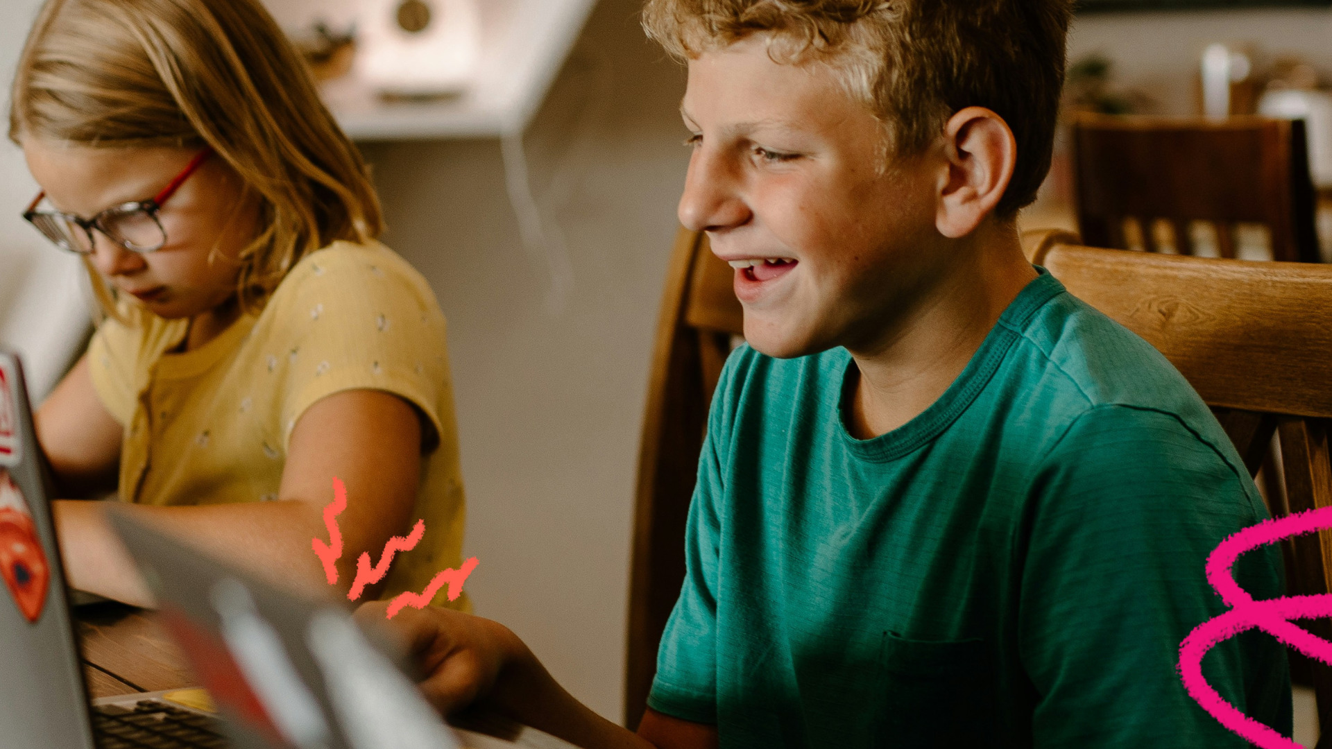 Imagem mostra um menino branco, de cabelo loiro curto e camisa verde sentando ao lado de uma menina branca de cabelos lisos e óculos. Eles estão usando computadores.