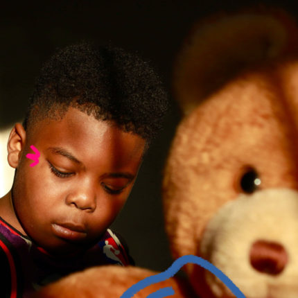 Imagem de capa para matéria sobre racismo na escola mostra um menino negro e riscos coloridos perto dos olhos. Ele está ao lado de um urso de pelúcia marrom.