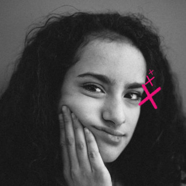 Imagem mostra retrato em preto e branco de uma garota com cabelo cacheado, expressão pensativa e leve sorriso. Ela apoia o rosto em uma das mãos, enquanto um desenho digital em rosa, em forma de “X” com pequenos traços, aparece próximo ao seu olho direito. O fundo é neutro. Imagem ilustra reportagem sobre a hora certa do primeiro celular na infância.