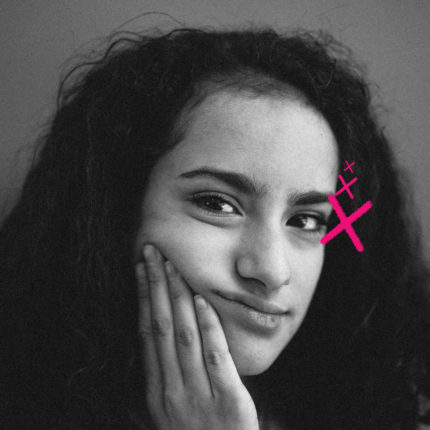 Imagem mostra retrato em preto e branco de uma garota com cabelo cacheado, expressão pensativa e leve sorriso. Ela apoia o rosto em uma das mãos, enquanto um desenho digital em rosa, em forma de “X” com pequenos traços, aparece próximo ao seu olho direito. O fundo é neutro. Imagem ilustra reportagem sobre a hora certa do primeiro celular na infância.