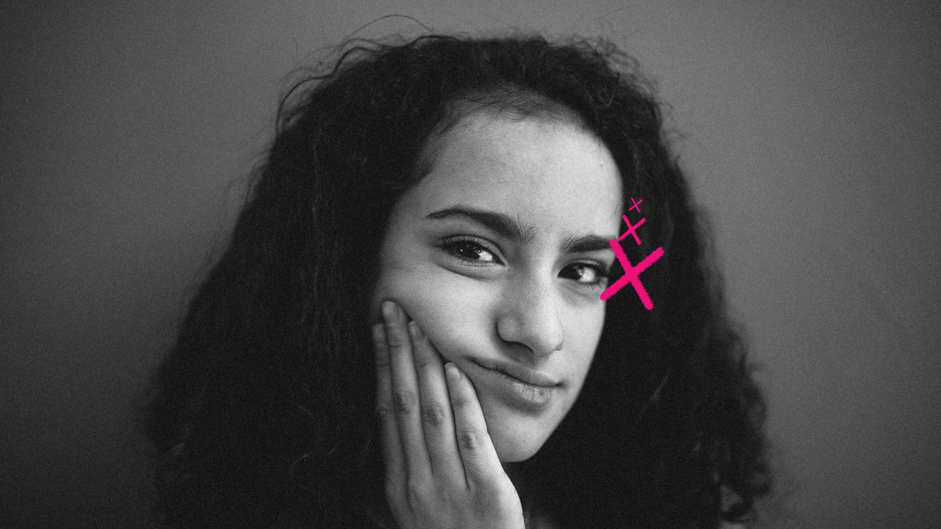 Imagem mostra retrato em preto e branco de uma garota com cabelo cacheado, expressão pensativa e leve sorriso. Ela apoia o rosto em uma das mãos, enquanto um desenho digital em rosa, em forma de “X” com pequenos traços, aparece próximo ao seu olho direito. O fundo é neutro. Imagem ilustra reportagem sobre a hora certa do primeiro celular na infância.