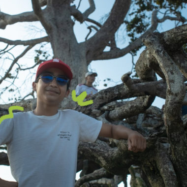 Imagem mostra um adolescente pardo usando camisa branca e boné vermelho ao lado de uma raiz de árvore.
