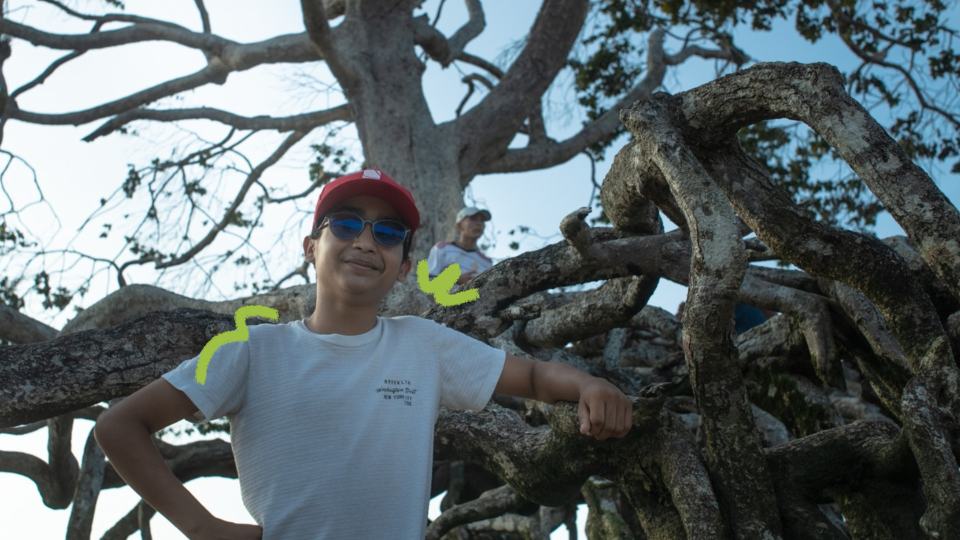 Imagem mostra um adolescente pardo usando camisa branca e boné vermelho ao lado de uma raiz de árvore.