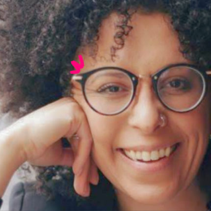 Imagem mostra foto de Natália Sousa, uma mulher negra de cabelos cacheados, usando óculos e sorrindo para a câmera. Ela fala sobre representatividade na infância.
