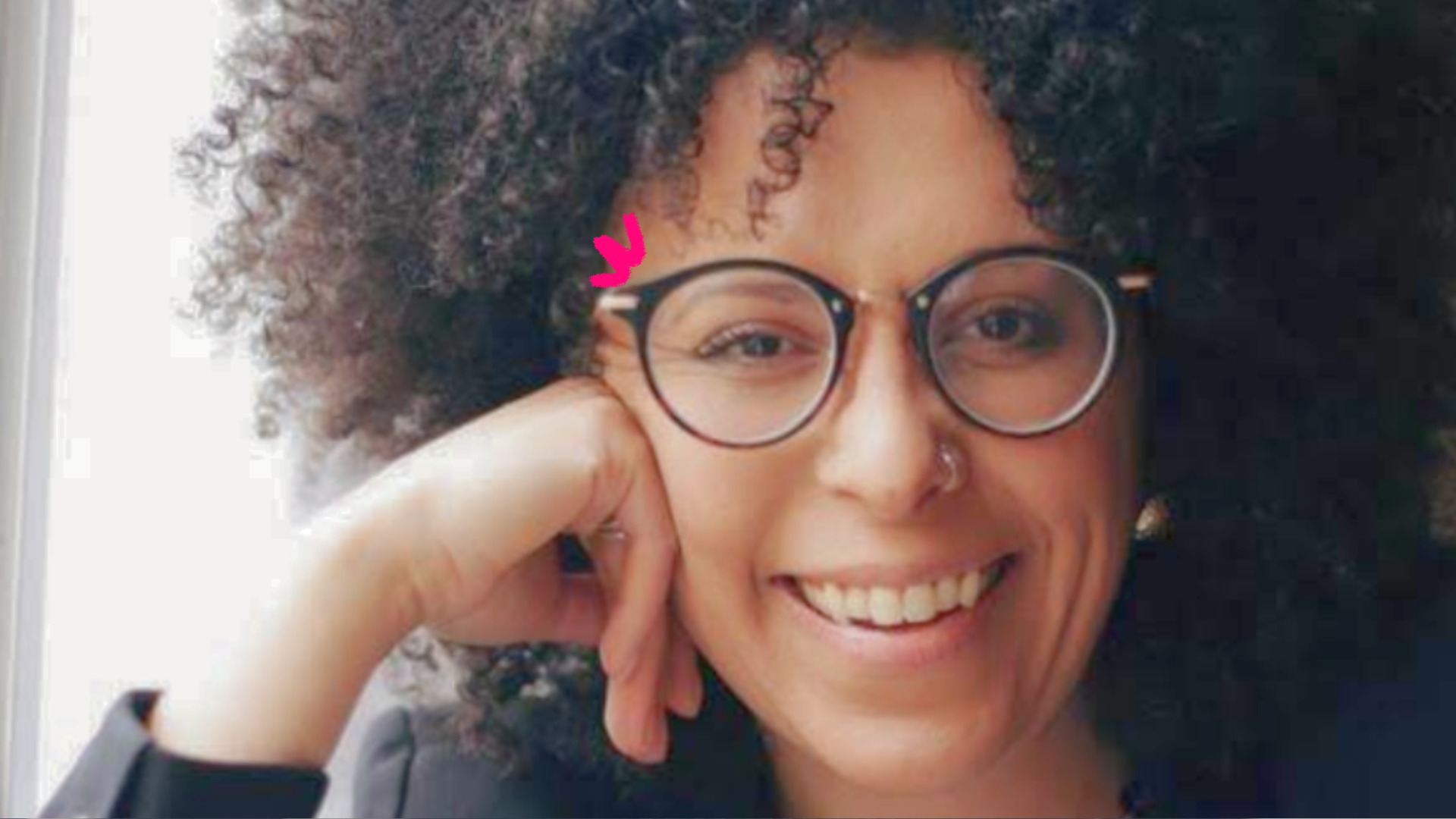 Imagem mostra foto de Natália Sousa, uma mulher negra de cabelos cacheados, usando óculos e sorrindo para a câmera. Ela fala sobre representatividade na infância.