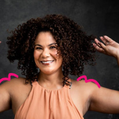 Imagem mostra Ediane Ribeiro, uma mulher negra, sorrindo e com os braços levantados em uma pose descontraída. Ela tem cabelos cacheados volumosos e veste uma blusa sem mangas em tom pêssego. Ela posa em um fundo escuro. Foto para falar sobre representatividade na infância.