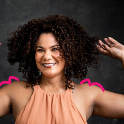 Imagem mostra Ediane Ribeiro, uma mulher negra, sorrindo e com os braços levantados em uma pose descontraída. Ela tem cabelos cacheados volumosos e veste uma blusa sem mangas em tom pêssego. Ela posa em um fundo escuro. Foto para falar sobre representatividade na infância.