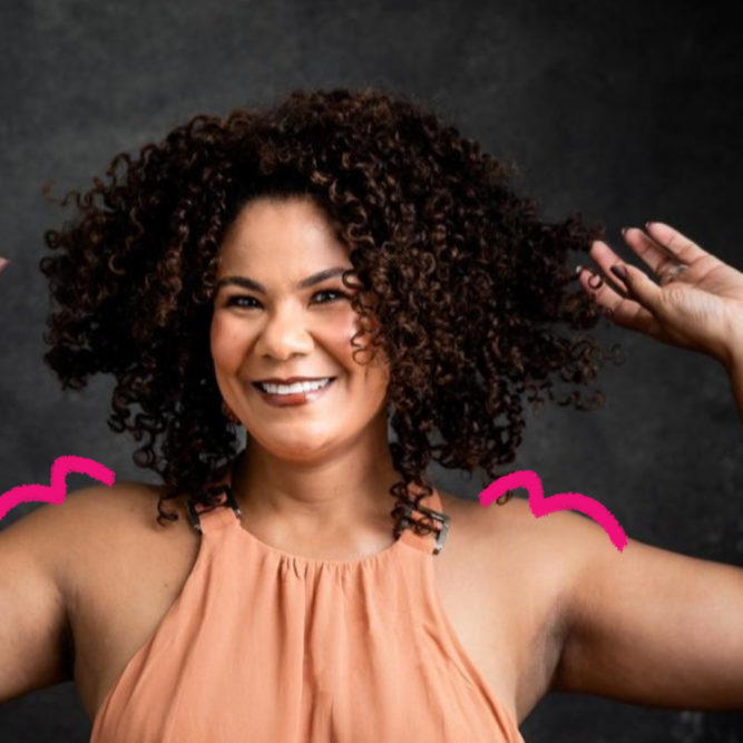 Imagem mostra Ediane Ribeiro, uma mulher negra, sorrindo e com os braços levantados em uma pose descontraída. Ela tem cabelos cacheados volumosos e veste uma blusa sem mangas em tom pêssego. Ela posa em um fundo escuro. Foto para falar sobre representatividade na infância.