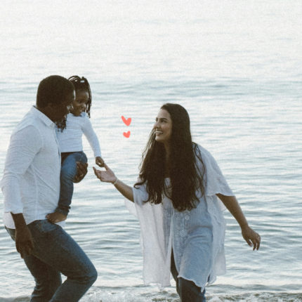 Imagem mostra um homem e uma mulher em uma praia e uma criança no colo do homem.