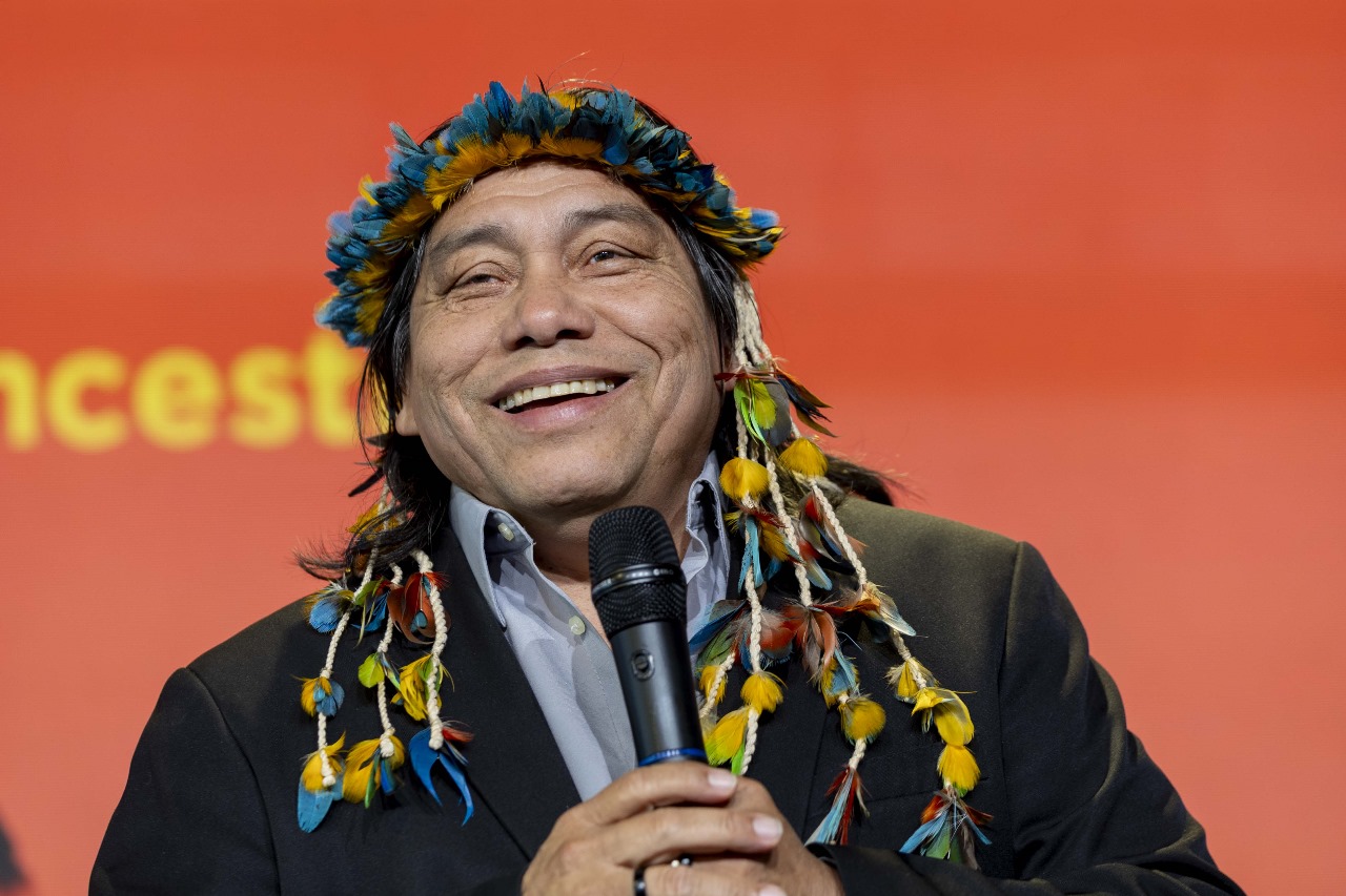 Imagem mostra foto do escritor indígena Daniel Munduruku. Ele sorri enquanto segura um microfone durante uma apresentação. Ele usa um cocar de penas coloridas e veste blazer escuro sobre camisa clara.