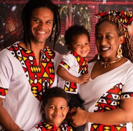 Imagem mostra foto da família da pedagoga Tatiane Santos. Ela, seu marido e os dois filhos pequenos estão sorridentes em frente a um cenário natalino. Todos usam roupas brancas com estampas geométricas coloridas de inspiração africana. Ao fundo, há decoração com árvore de Natal e luzes.