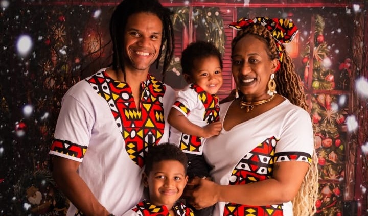 Imagem mostra foto da família da pedagoga Tatiane Santos. Ela, seu marido e os dois filhos pequenos estão sorridentes em frente a um cenário natalino. Todos usam roupas brancas com estampas geométricas coloridas de inspiração africana. Ao fundo, há decoração com árvore de Natal e luzes.