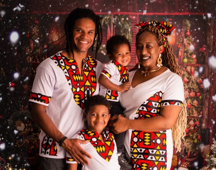 Imagem mostra foto da família da pedagoga Tatiane Santos. Ela, seu marido e os dois filhos pequenos estão sorridentes em frente a um cenário natalino. Todos usam roupas brancas com estampas geométricas coloridas de inspiração africana. Ao fundo, há decoração com árvore de Natal e luzes.