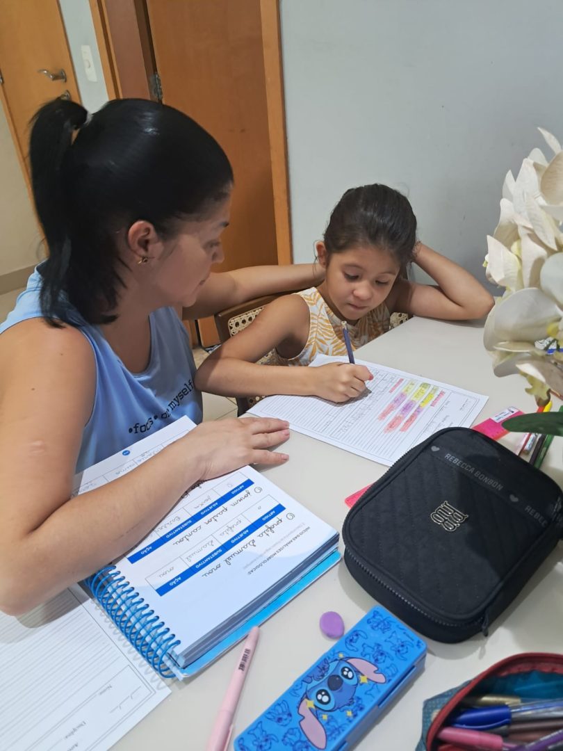 Imagem mostra Daniele, uma mulher branca de cabelo preto liso, sentada à mesa ao lado de Katarina, uma menina branca, de cabelo liso preto. As duas estão estudando com livros e canetas na mesa.