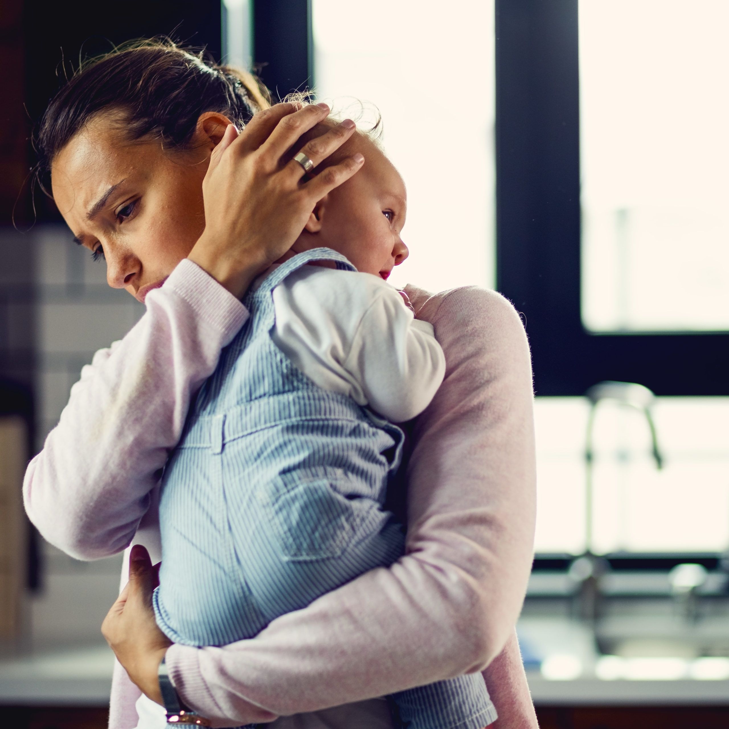 Mãe segura o bebê no colo em um abraço apertado, apoiando a cabeça da criança no ombro, em um ambiente doméstico iluminado por luz natural. A expressão da mãe revela emoção e apreensão, simbolizando o momento de separação e os sentimentos envolvidos no primeiro dia de creche, como cuidado, vínculo e adaptação.