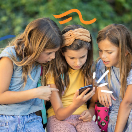 Três meninas olham para a tela de um celular nas mãos da menina do meio. Todas são brancas e vestem roupas coloridas.