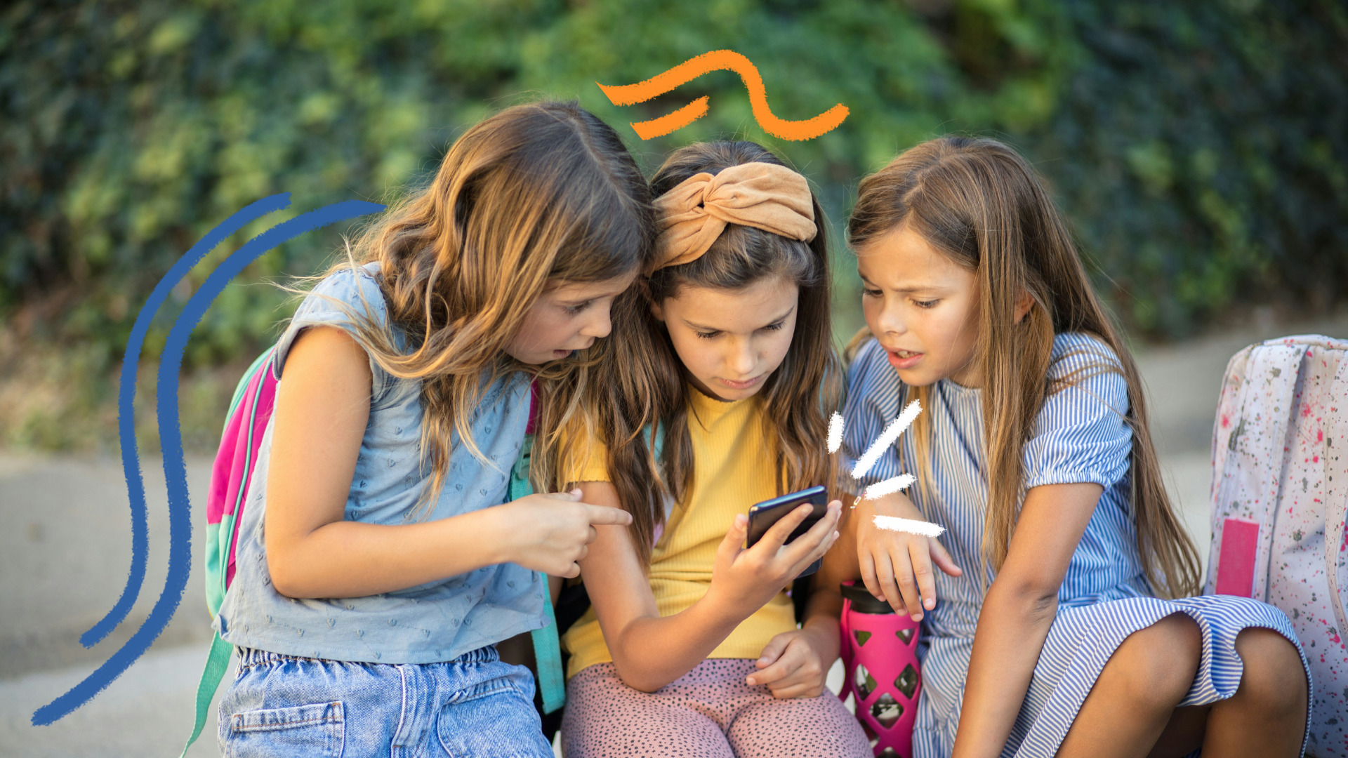 Três meninas olham para a tela de um celular nas mãos da menina do meio. Todas são brancas e vestem roupas coloridas.