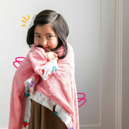 Uma menina de cabelo escuro está enrolada em uma mantinha cor de rosa em frente a uma porta branca