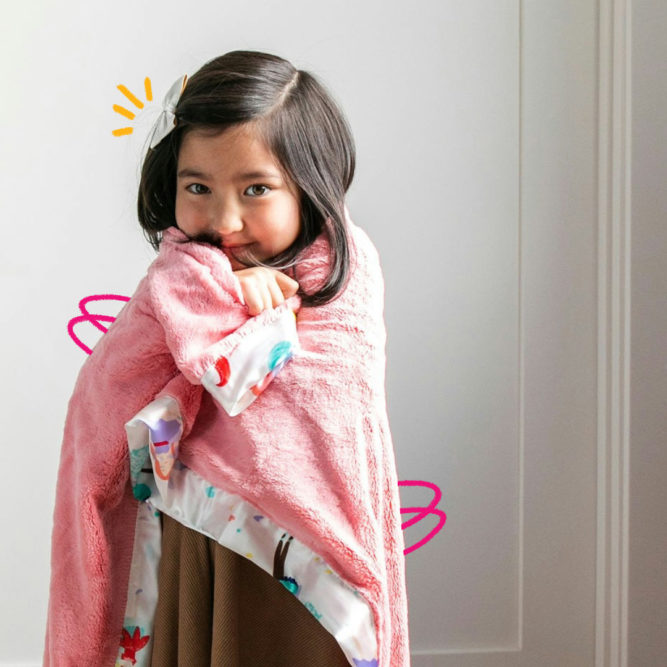 Uma menina de cabelo escuro está enrolada em uma mantinha cor de rosa em frente a uma porta branca