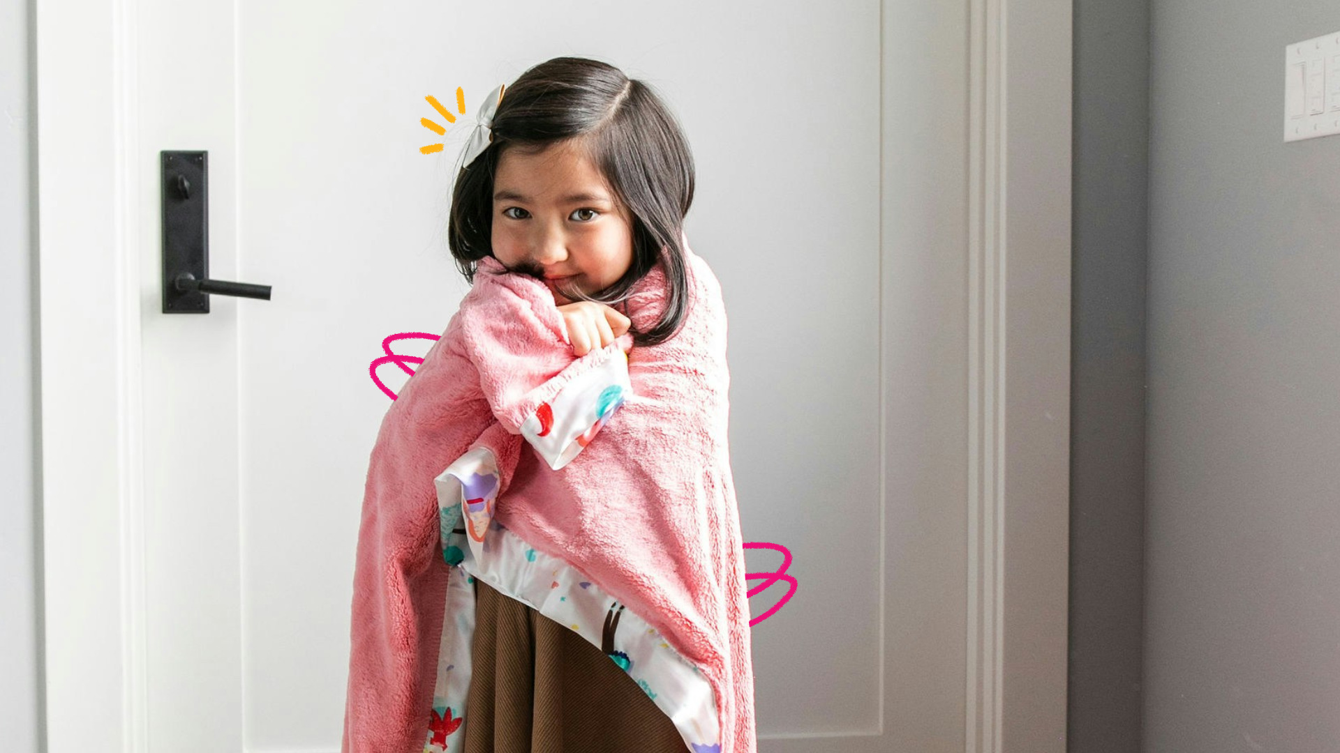 Uma menina de cabelo escuro está enrolada em uma mantinha cor de rosa em frente a uma porta branca
