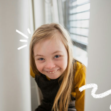 Menina com síndrome de Down sorri enquanto espia por entre uma cortina branca, vestindo blusa amarela e moletom escuro.